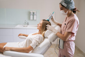 Side view of trichologist in gloves and mask performing hair treatment procedure using spectrum phototherapy device. Blonde female patient having hair light therapy